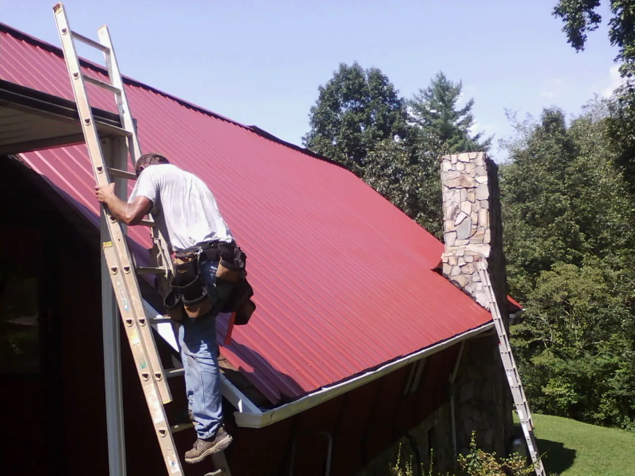 Roofer on a ladder beside a red metal roof and stone chimney — Soffit & Fascia Repair by Roofing Brothers in New Salem